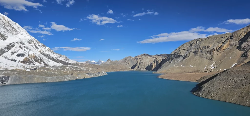 Vast, serene turquoise Tilicho Lake stretches out under a bright blue sky dotted with fluffy white clouds. The lake is nestled high in a rugged, mountainous landscape. To the left, a snow-capped mountain rises dramatically, its lower slopes meeting the rocky shoreline. On the right, barren, rocky mountains in shades of grey and brown slope down to the water's edge. The distant horizon reveals more jagged peaks under the clear sky, emphasizing the remoteness and high altitude of the location.